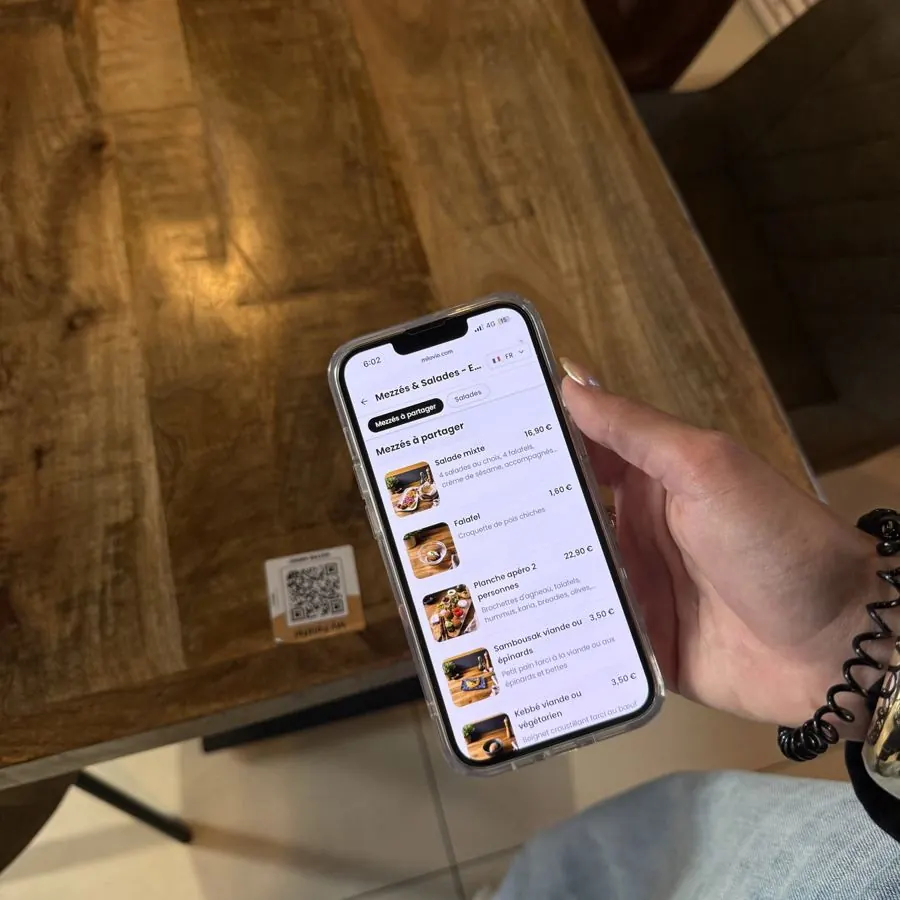 La carte des mezzés s’affiche sur un téléphone tenu au-dessus d’une table de My Falafel, avec l’Autocollant QR en arrière-plan.