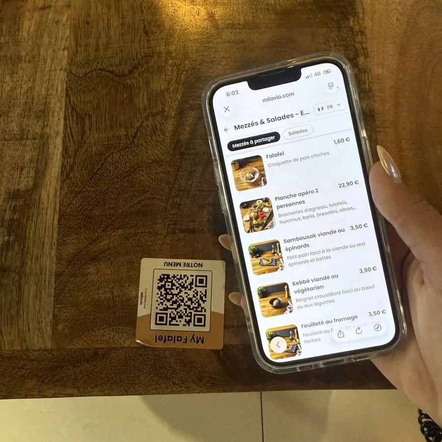Une main tient un téléphone ouvert sur la carte des mezzés de My Falafel, où le falafel s’affiche à 1,60 €, au-dessus de l’Autocollant QR de la table.